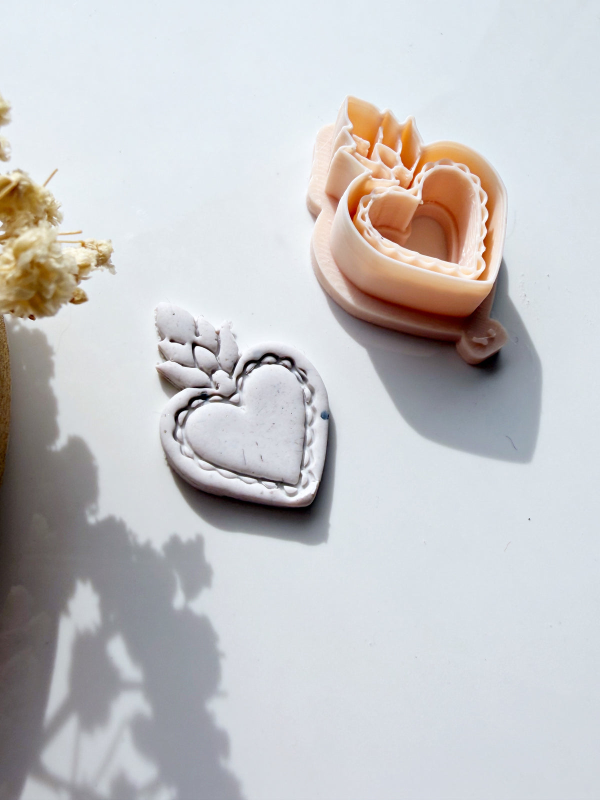 A Sacred Heart cookie cutter and a finished grey clay heart shape on a white surface, perfect for jewelry making.