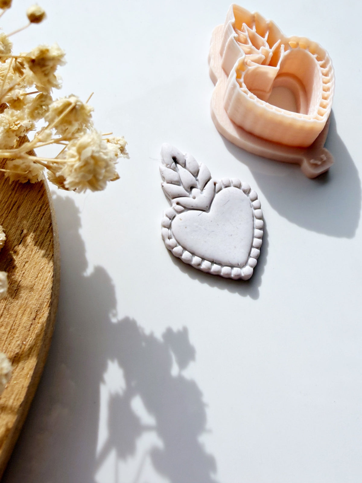 Sacred heart shaped clay cutter and a finished grey clay charm on a white background with dried flowers