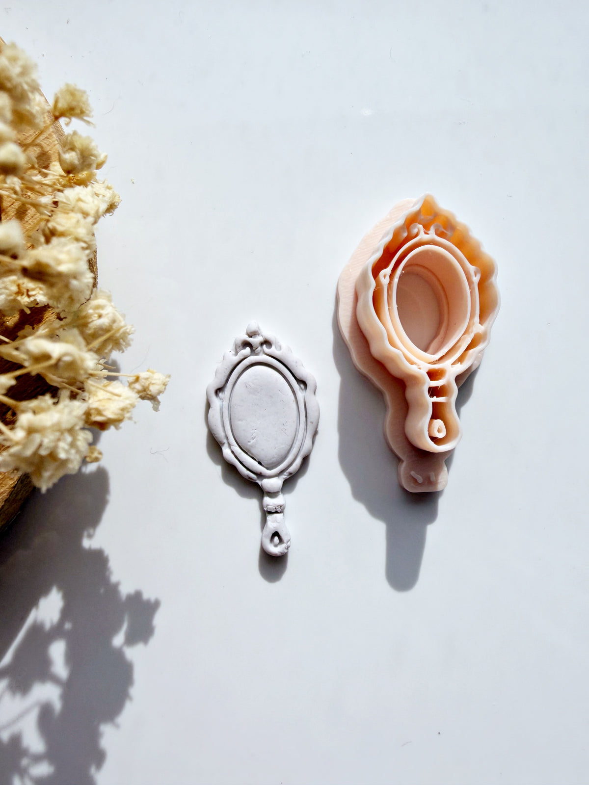Victorian mirror cookie cutter and fondant stamp with a clay impression on a white surface next to dried flowers.