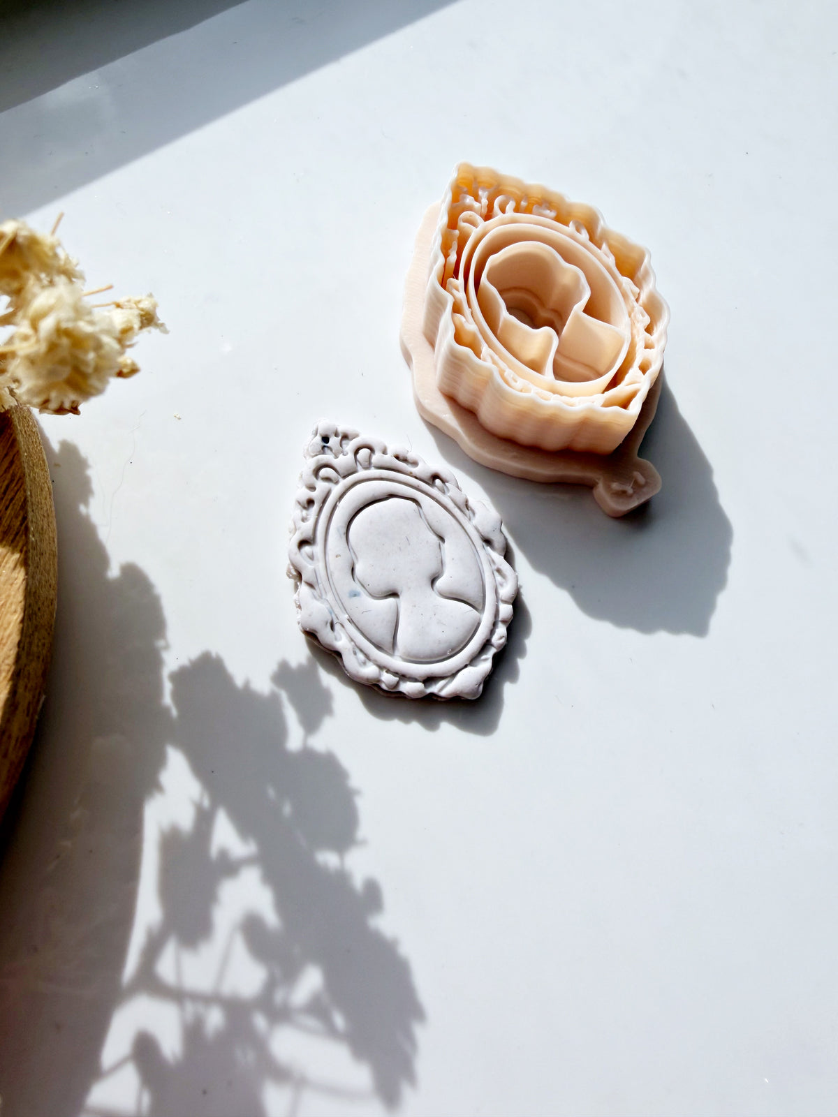 Cameo silhouette polymer clay cutter and finished white ornate frame pendant on a white surface.