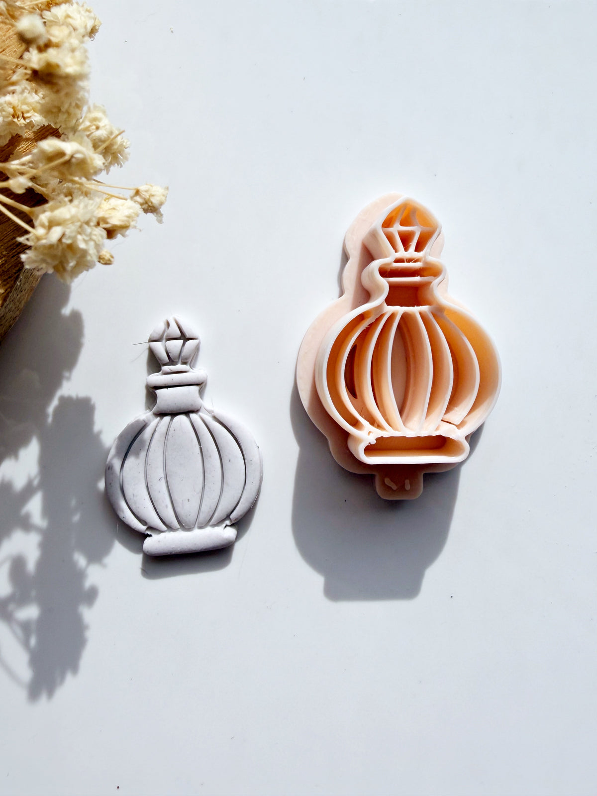 Perfume bottle cookie cutter and its corresponding clay shape on a white background with dried flowers.